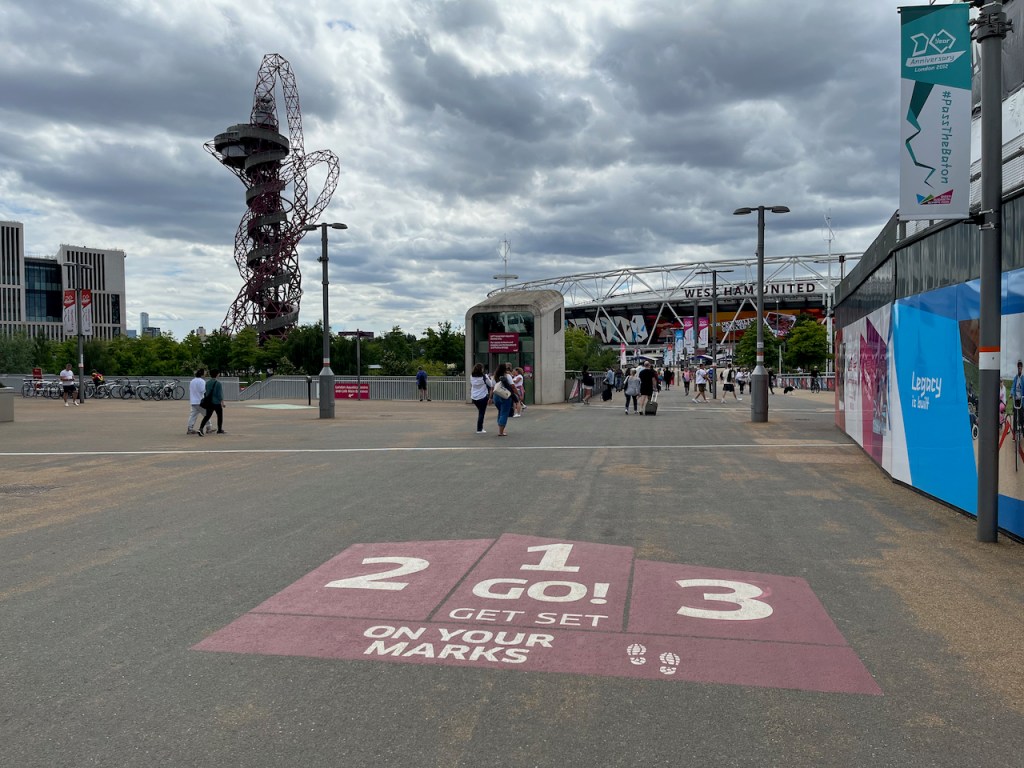Large pedestrian area leading into the Olympic Park, with the tall Orbit Tower and West Ham Stadium ahead. The shape of a medal podium in a dark shade of pink is painted on the pavement. Beneath it, as you walk towards it, white letter says On Your Marks, Get Set. Then the numbers 3, 2, 1, on their respective podium positions, sit around the word Go.