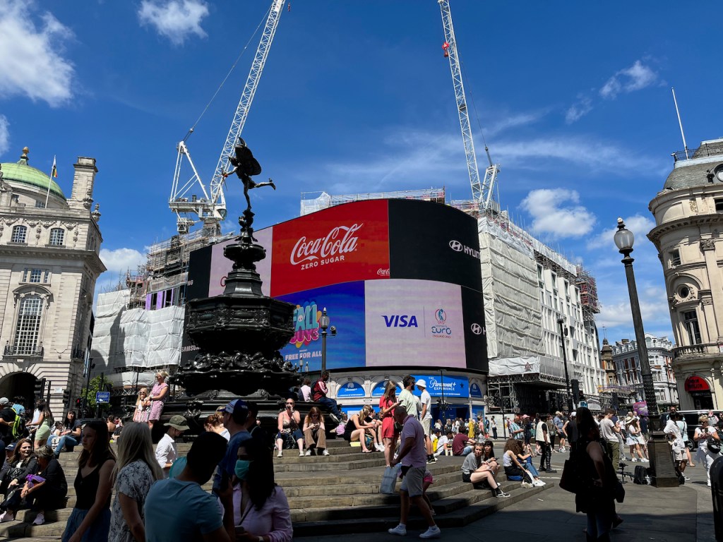 Piccadilly Circus, on a busy day in the sunshine. The tall, black, ornate Shaftesbury Memorial Fountain stands in the centre, and perched on one leg on top of it is a statue of a nude man with large wings, firing a bow and arrow. Behind it are the big Piccadilly Circus video billboards, including adverts for Coca-Cola and Visa.