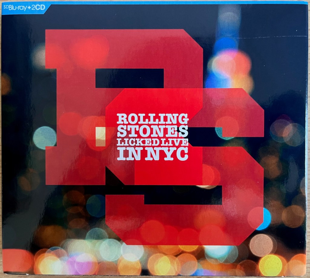 Front cover for The Rolling Stones, Licked Live In NYC. The title is in white text over 2 large overlapping red initials R and S. The background is a scene of blurry lights.