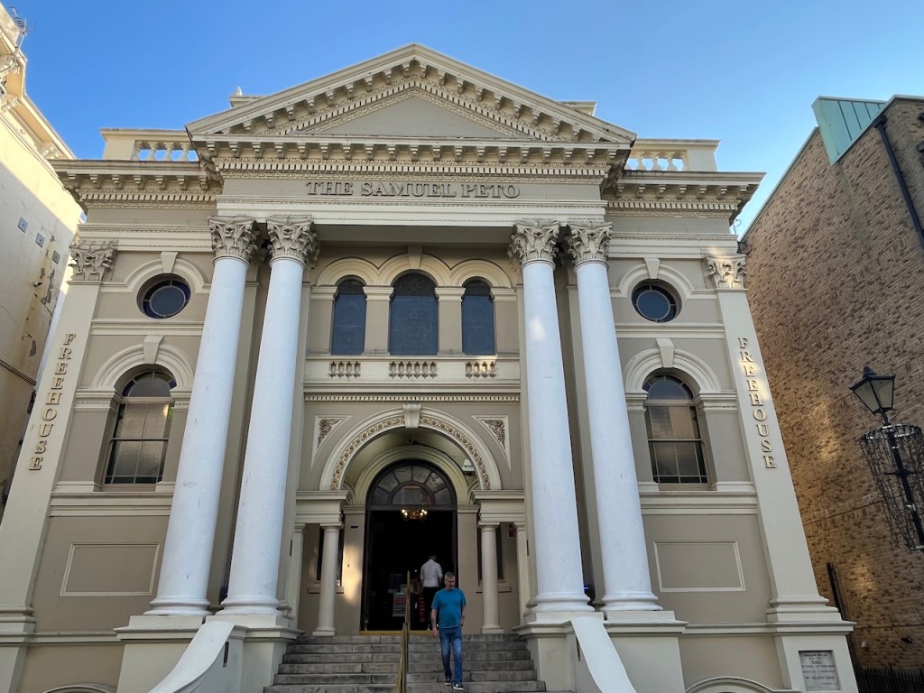 The exterior of The Samuel Peto, a Wetherspoons pub in a converted chapel. The white building is quite ornate, with pairs of 2-storey high white pillars either side of the arched doorway, and a triangular roof section above.