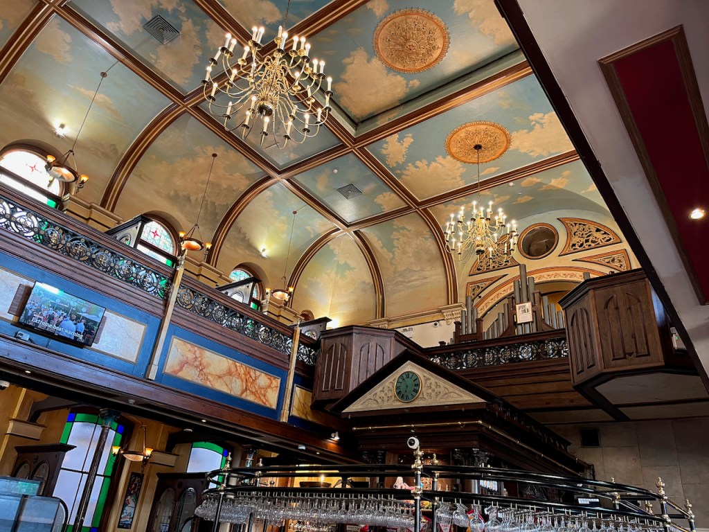 The ornate interior of The Samuel Peto, the Wetherspoons pub in a converted chapel. The ceiling panels are designed to represent a blue sky with a few white clouds, hanging from which are a couple of ornate chandeliers. In the background, the pipes of the chapel organ are visible on the upper level. To the left, a TV mounted above the ground floor is showing the BBC News, with the headline Extreme Heat Warning.