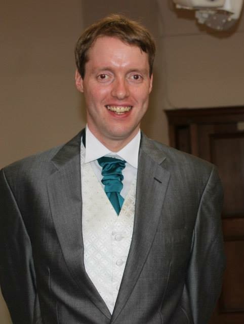 Glen smiling in church while dressed in his grey best man suit with white shirt and a green ruffled tie.