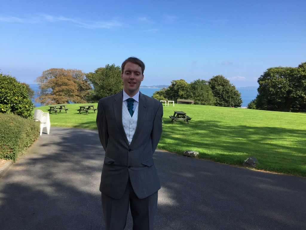 Glen wearing a grey suit with a white shirt, standing in front of a sunny grassy area with benches and a sea view, beneath a blue sky.