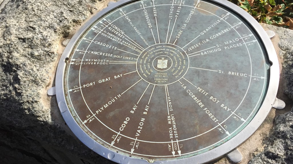 Large round plaque with arrows pointing in all directions from the centre towards the outer edge, each marked with the distance to a different place in Guernsey, the UK, Europe and beyond. Text around a coat of arms in the centre reads Their majesties King George & Queen Mary, accompanied by Princess Mary on board the royal yacht, arrived in the roadstead on Sunday July 10th 1921, and visited all parts of the island on the following day. Latitude 49, 27, 40 north. Longitude 2, 32, 15 west.