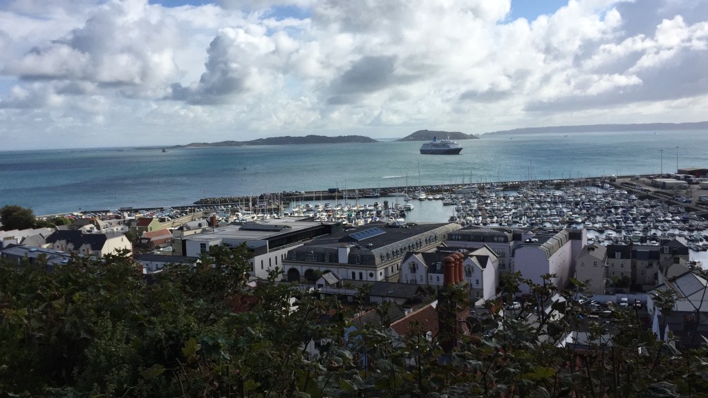 View of Guernsey harbour in the distance, with a large ferry in the open water beyond it, and the island of Herm visible on the horizon.
