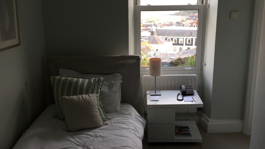 Bedroom in La Fregate Hotel in Guernsey. A single bed with a couple of cushions in front of the pillows is next to a bedside table positioned in front of the window. The table has a lamp and a telephone, plus a small pad and a pen. The rooftops of nearby buildings and the sea in the distance can be seen through the window.