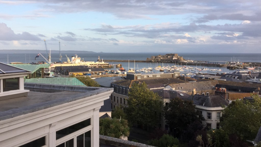 View from La Fregate Hotel towards Guernsey harbour in the sunshine, with lots of boats moored up. At the far side of the harbour is the long breakwater walk leading to a lighthouse.