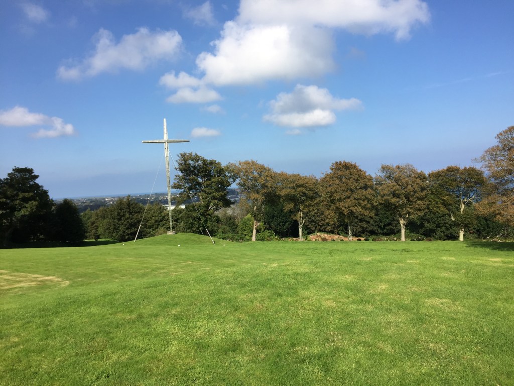 Large area of grass in the sunshine. In front of the trees lining the edge is a very tall pointed metal cross.