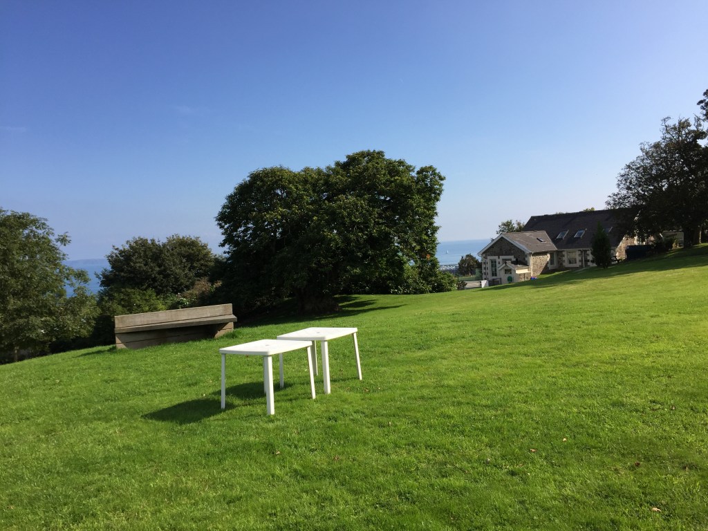 Large area of grass in the sunshine, with a concrete bench and a couple of small square white tables. Trees line the edge, with a space revealing the upper floor of a house, while the sea is just visible in the far distance behind it.