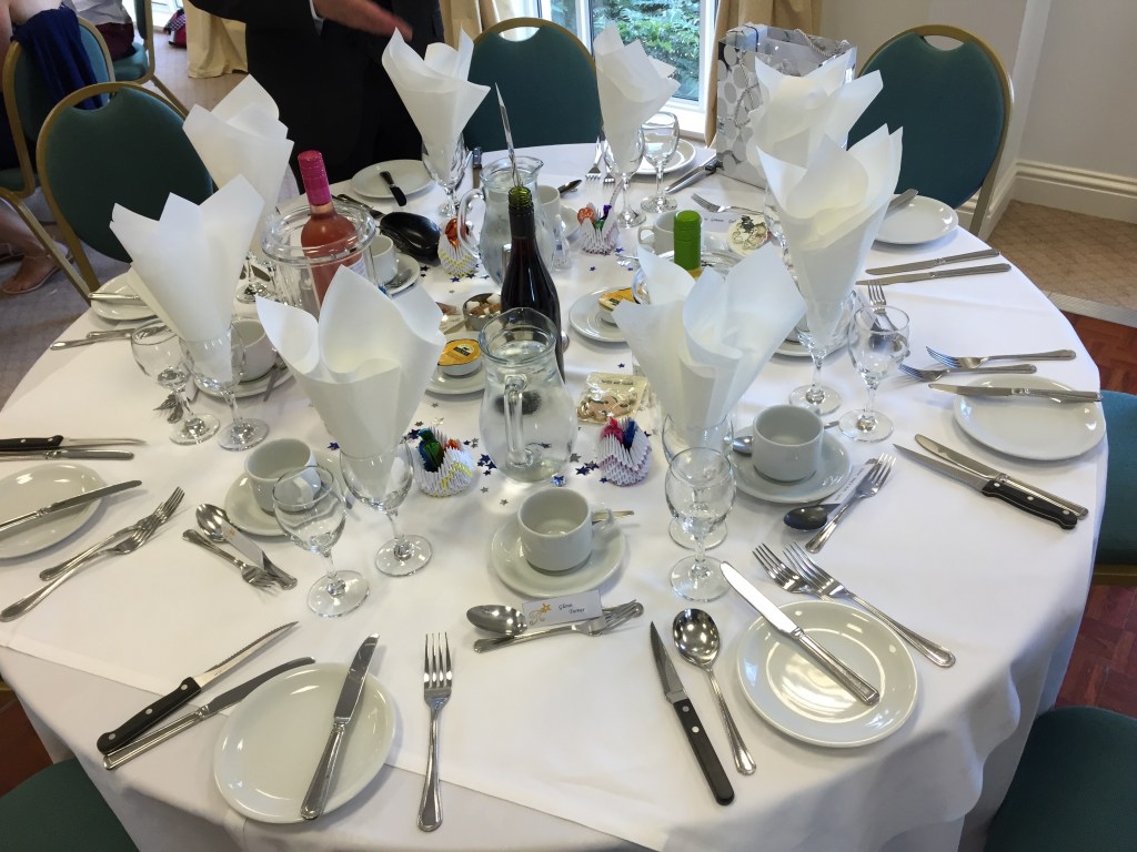 A round table set up for a wedding reception dinner. Each named place at the table has cutlery laid out, a mug and saucer, an empty glass, and a second glass with a folded napkin inside it. The centre of the table has bottles of wine, little swans out of paper carrying complementary sweets, small pots of butter, and lots of little stars decorating the white tablecloth.