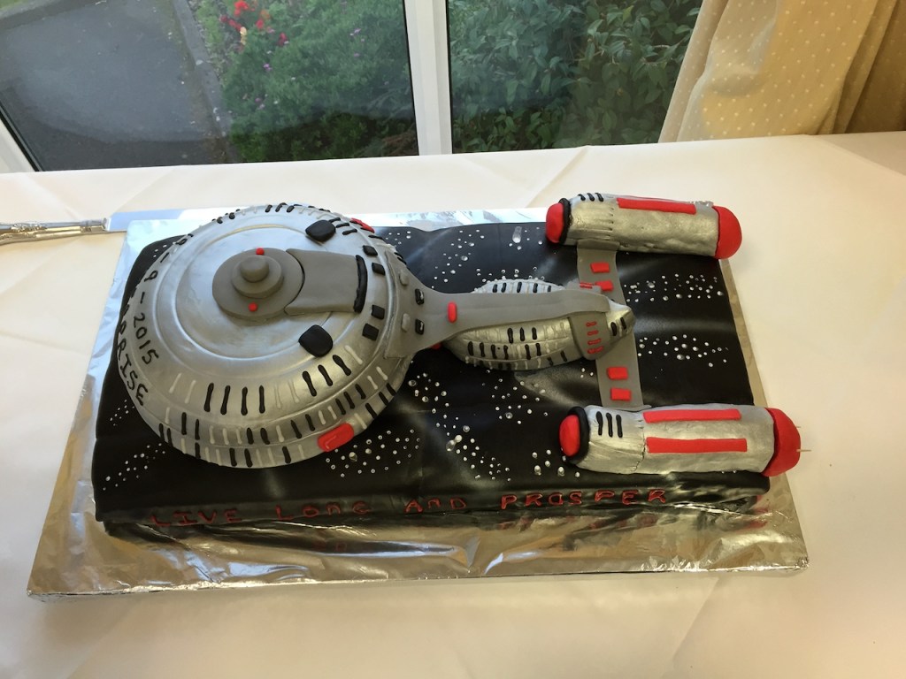 A large, detailed cake in the shape of the Starship Enterprise, with the grey ship on top of a base that is black and covered with white stars to look like space.