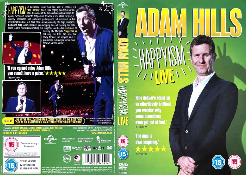 DVD cover spread for Adam Hills - Happyism. The front cover shows Adam standing proudly in a black jacket and a white shirt, while the back cover shows photos of him performing on stage, in one of which he has his mouth wide open in shock.