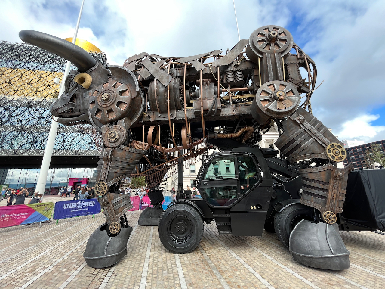Left side view of the very large animatronic bull from the Commonwealth Games, made up of lots of different metal parts including wheels and chains. A cabin with big wheels sits beneath the bull's chest, which can be used to drive it around.