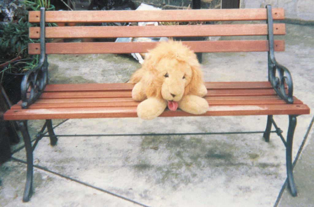 A cuddly toy lion sitting on a garden bench, with its tongue sticking out.