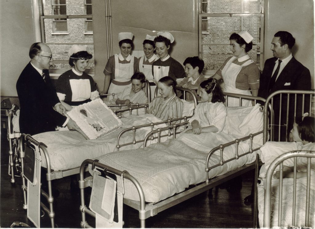 A black and white photo of 4 girls in hospital beds next to each other, with a fifth girl standing behind the central bed. They are surrounded by 5 female nurses and 2 gentlemen in suits. The girls are all smiling as they look at a large cake being held up for them to see by one of the nurses, which has the words "Greetings On Your 33rd Anniversary" on it, surrounded by floral, decorative icing.