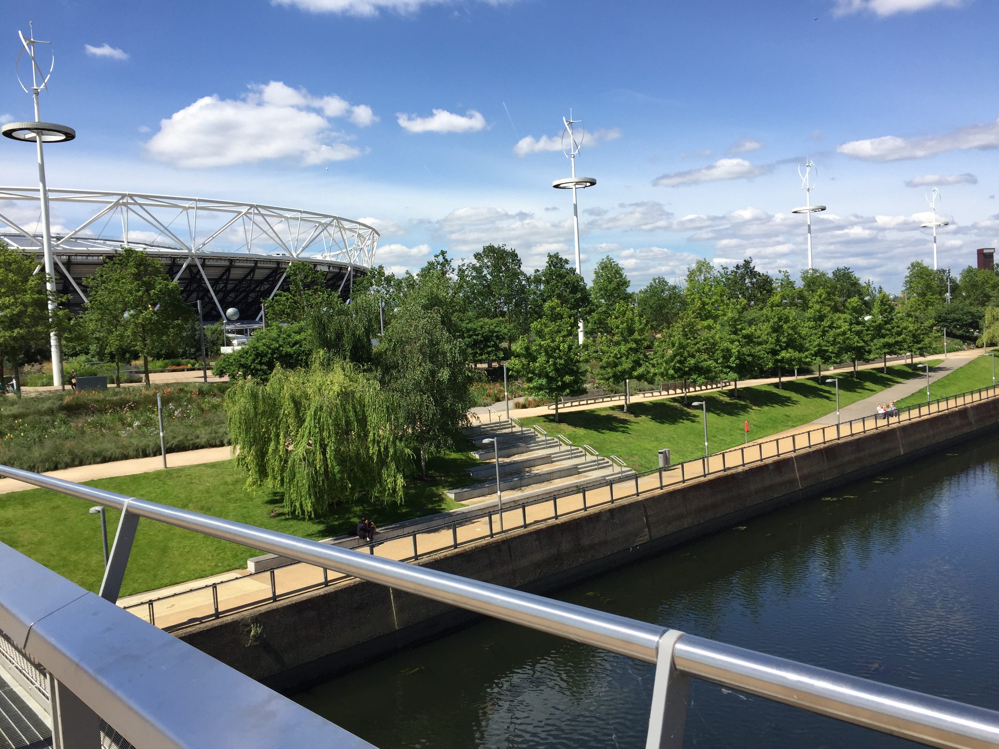 View towards the bank of a river flowing through the Olympic, with leafy trees, long patches of grass and footpaths, with the Olympic Stadium in the background.