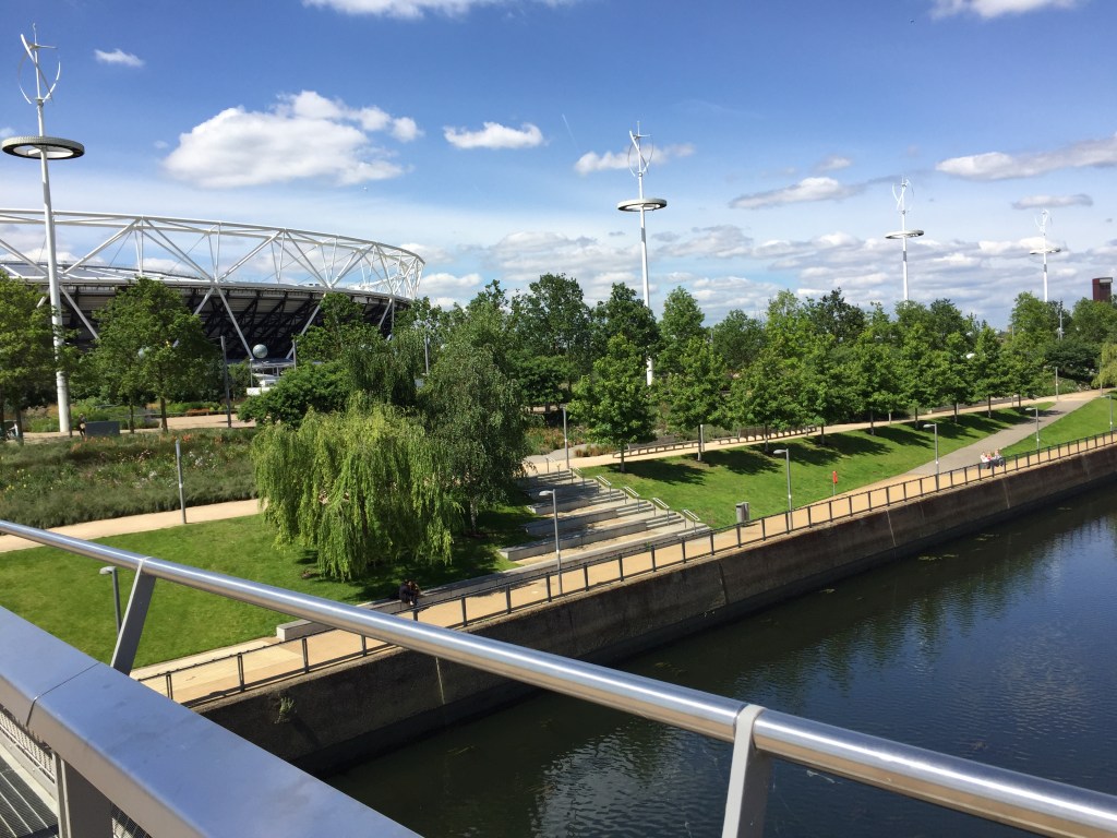View towards the bank of a river flowing through the Olympic, with leafy trees, long patches of grass and footpaths, with the Olympic Stadium in the background.