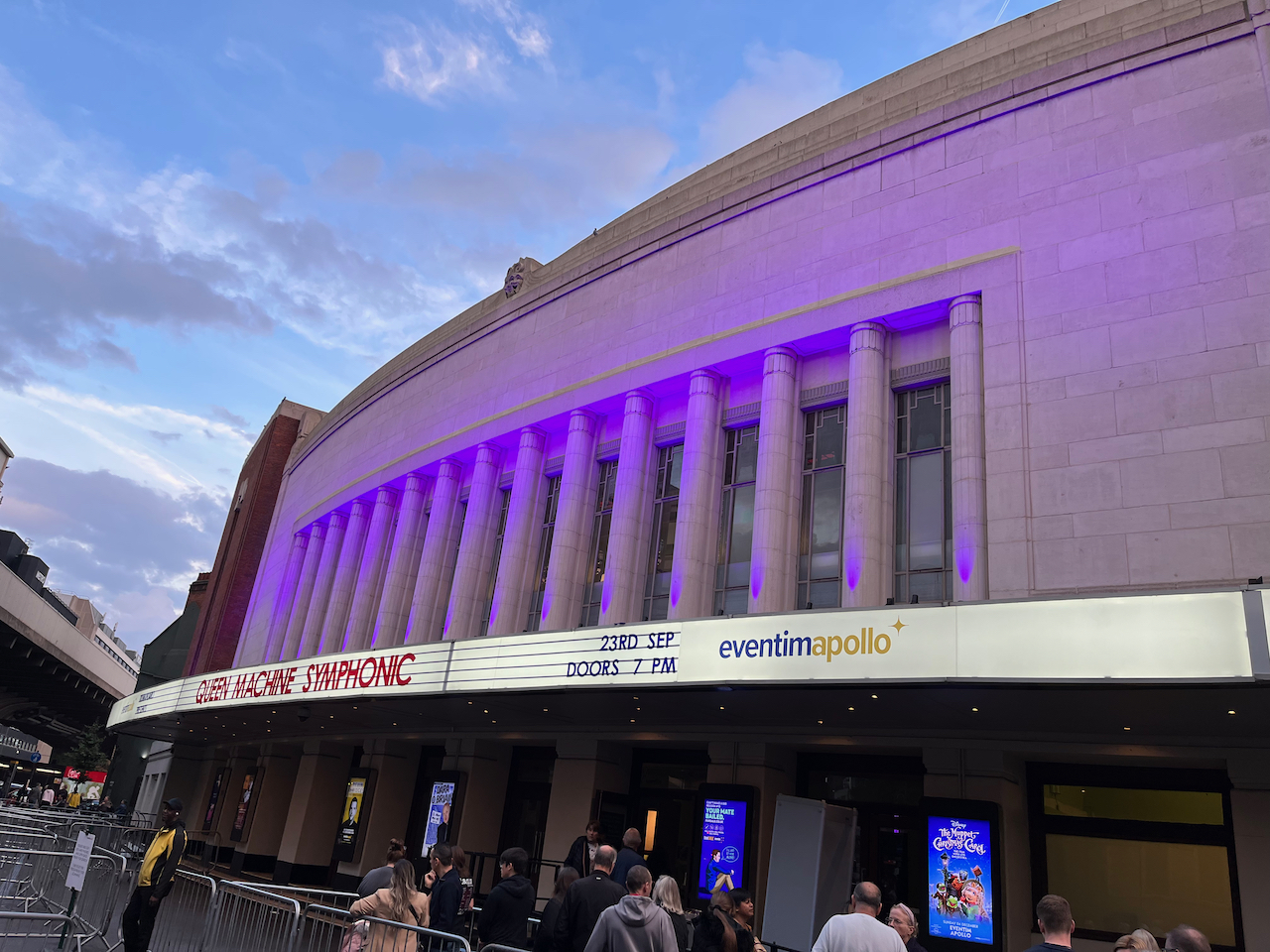 The Eventim Apollo building in Hammersmith, with writing on the long white lit-up curving canopy over the entrance doors that reads Queen Machine Symphonic, 23rd September, doors 7pm.