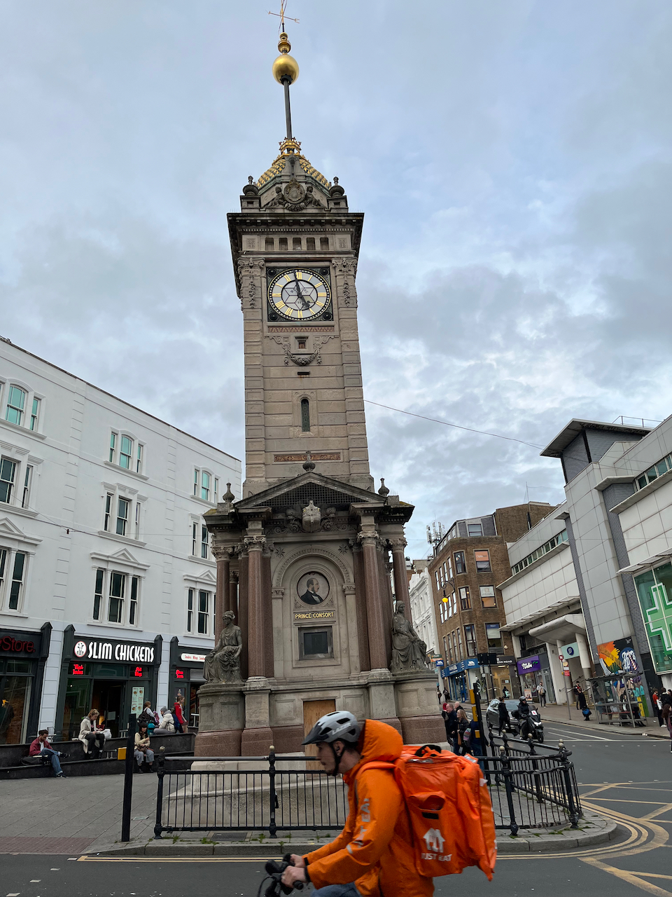 Tall, ornate clock tower in Brighton's shopping district. The clock face has a white background, black lines and hands, and gold roman numerals.
