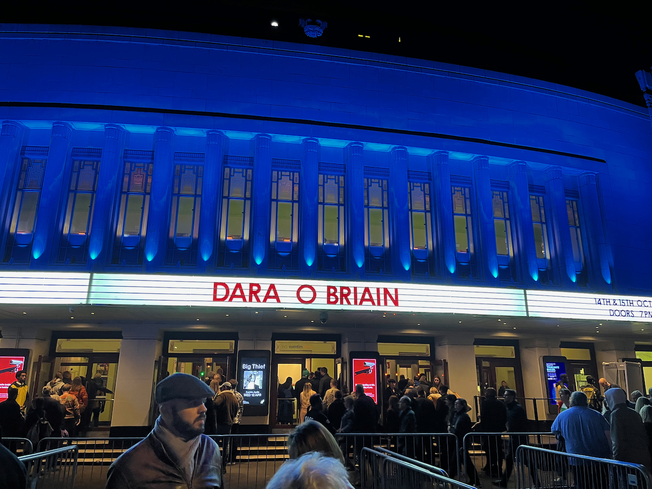 The Hammersmith Apollo lit up in blue, as a large crowd of people enter to see Dara O Briain, whose name is in big red letters on the white canopy over the doors.