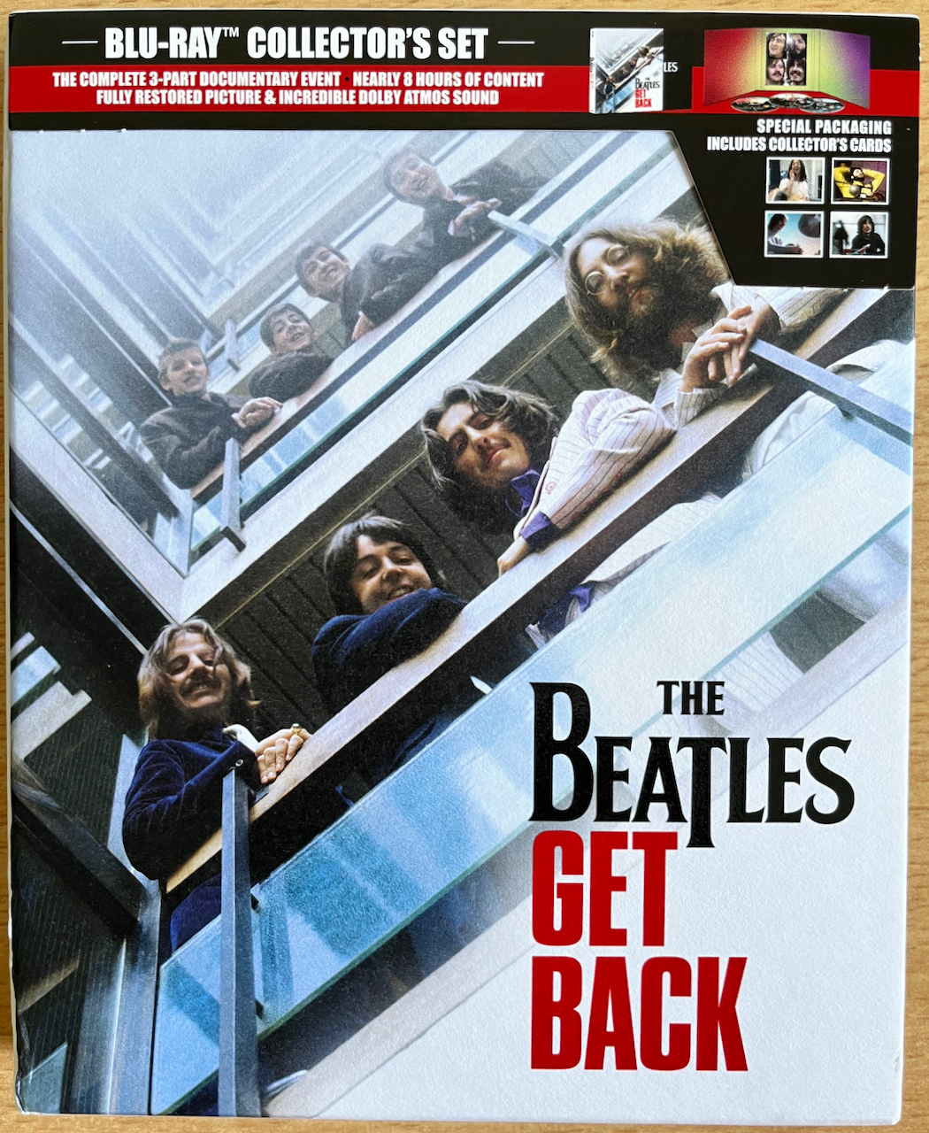 Front cover of the Blu-ray set for Get Back by The Beatles, showing the 4 band members smiling as they look over a railing in a tower block, with younger versions of themselves looking over a railing on the floor above.