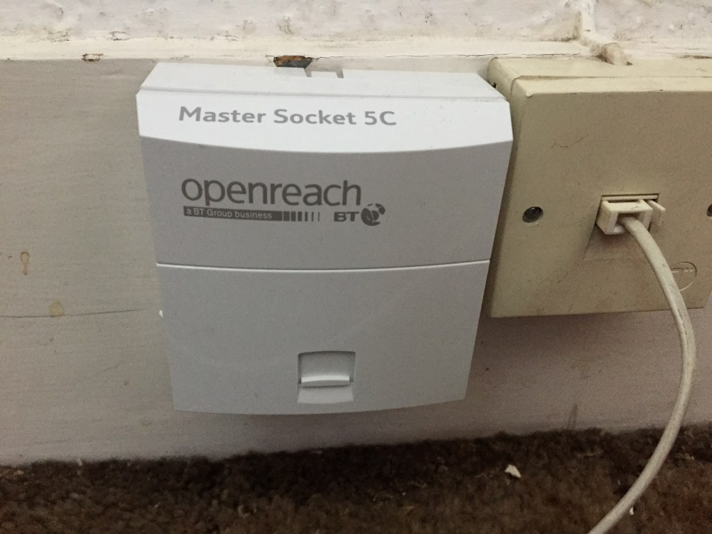 A small white phone socket box on a skirting board, with the BT Openreach logo on the front, and the text Master Socket 5C on the top. There is an older, smaller phone socket to the right of it.