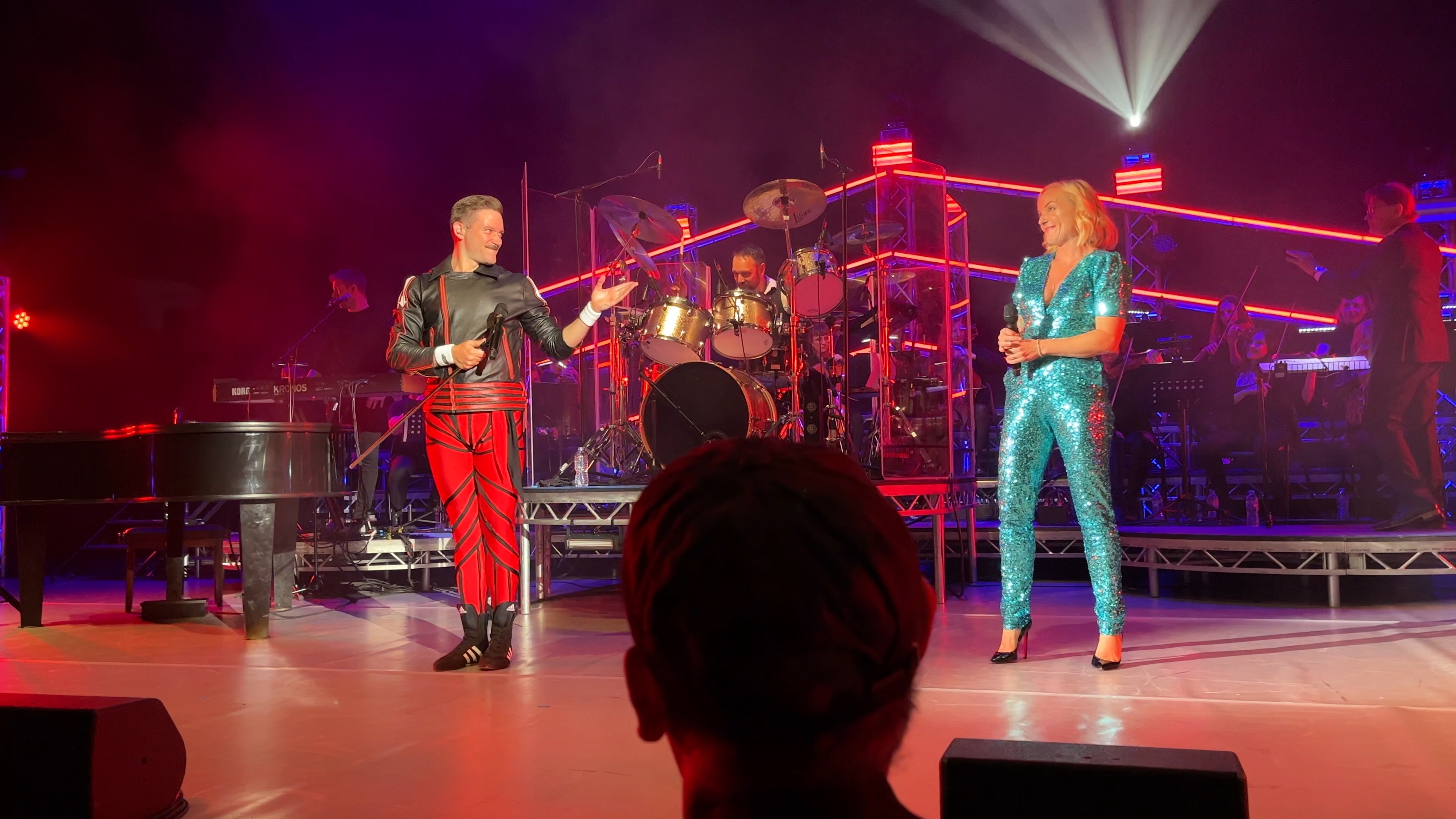 Queen Machine in concert at the Hammersmith Apollo. The lead singer looks a bit like Freddie Mercury, with his moustache and body-hugging outfit of black jacket and red trousers. He's smiling and gesturing towards West End star Kerry Ellis, a smiling blonde lady wearing a very sparkly, short-sleeved, low-cut, light blue bodysuit, along with black high heels. Behind them, to the right of the drummer on his large kit, the London Symphonic Rock Orchestra are playing on stringed instruments.