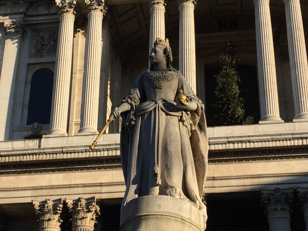 The ornate Queen Anne statue in front of the cathedral. She is wearing a long robe, the collar of the Order of the Garter, and a gilded crown. In one hand she is holding a gilded orb, and in the other a gilded sceptre.