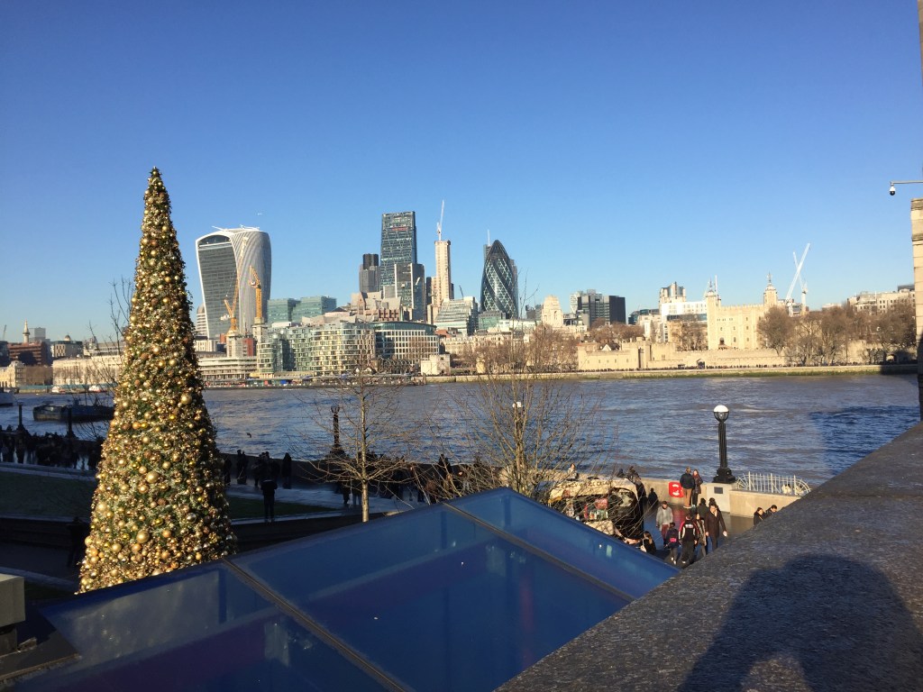 View from the South Bank of the Thames, where there is a tall, green, decorated Christmas tree, towards the Tower of London, Gherkin and Walkie-Talkie buildings on the opposite side of the river.