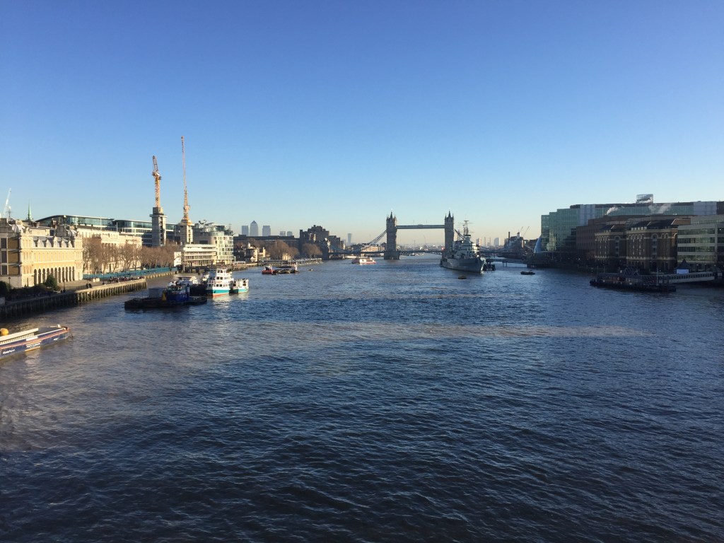 View along the Thames from London Bridge towards Tower Bridge, including the Tower of London on the left and HMS Belfast on the right, on a sunny day beneath a clear blue sky.