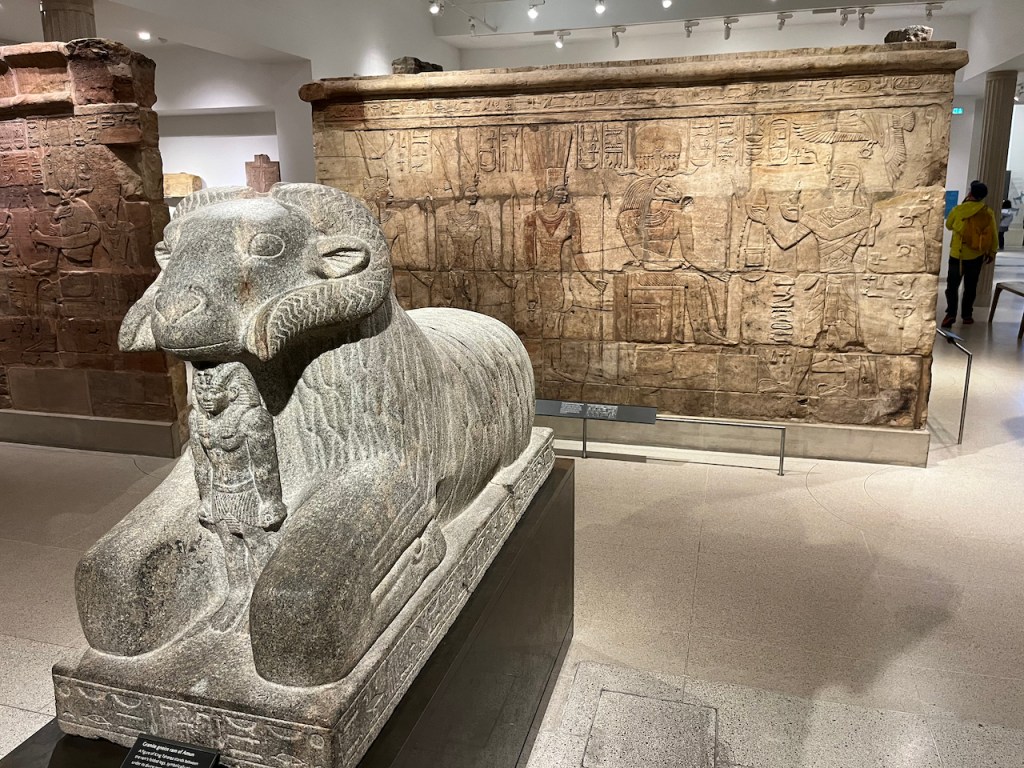 A statue of a ram, in front of a large wall engraved with images of people and hieroglyphics.