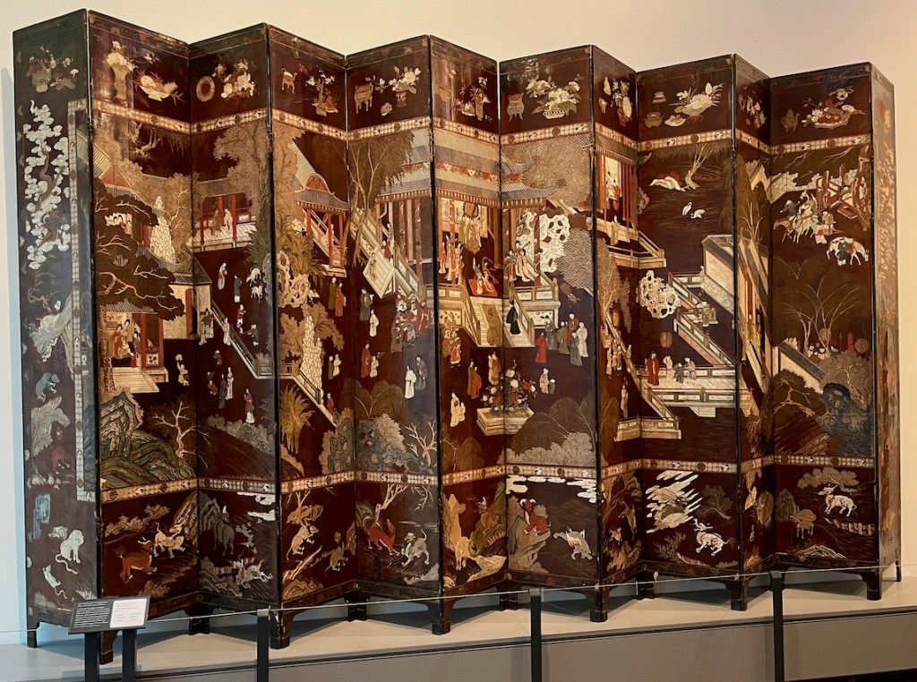 A large folding screen with a very detailed painting of a bustling town, including people, buildings, animals and plants.
