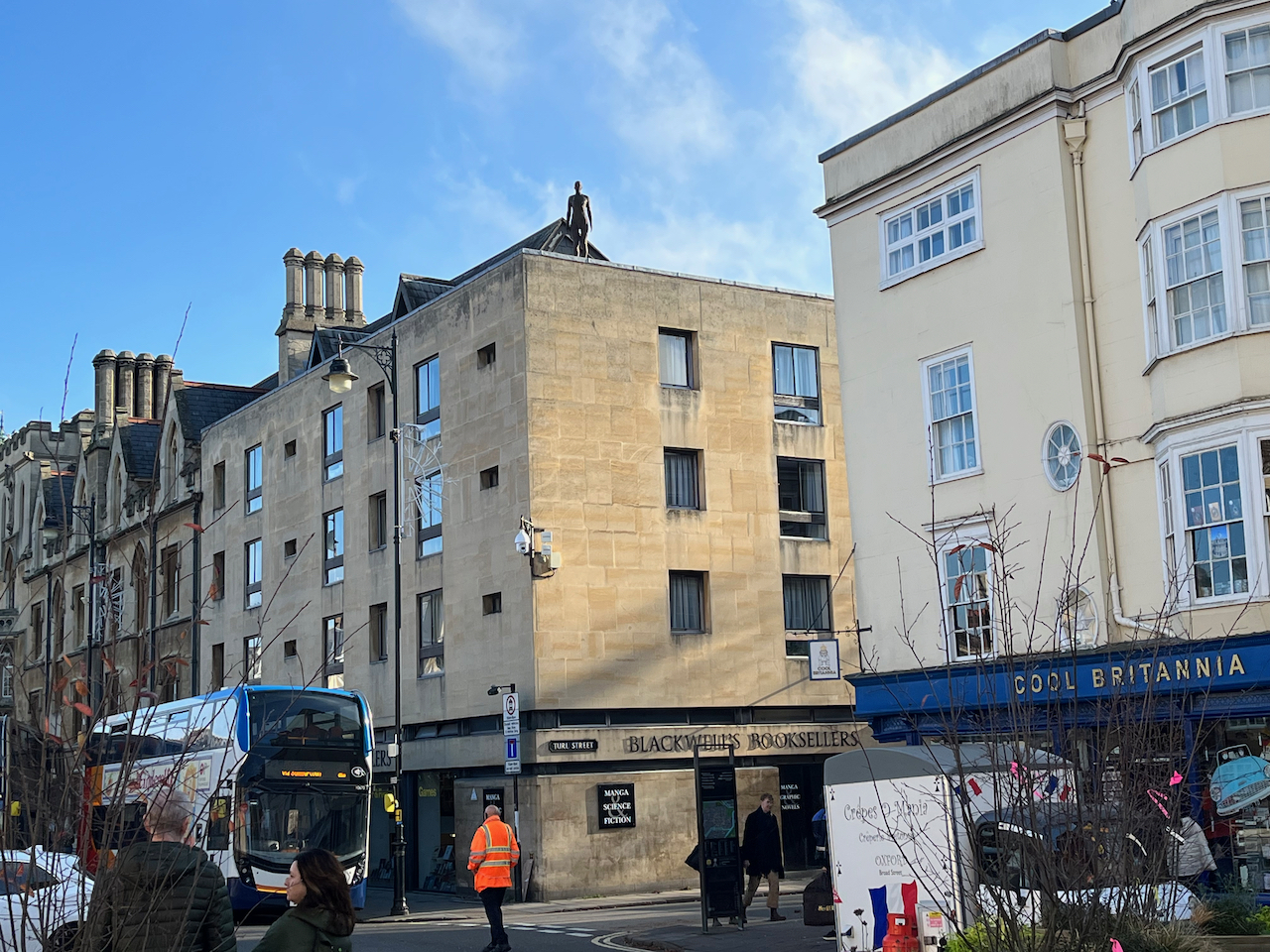 A plain black statue of a nude figure on the corner of the roof of Blackwell's Bookshop.