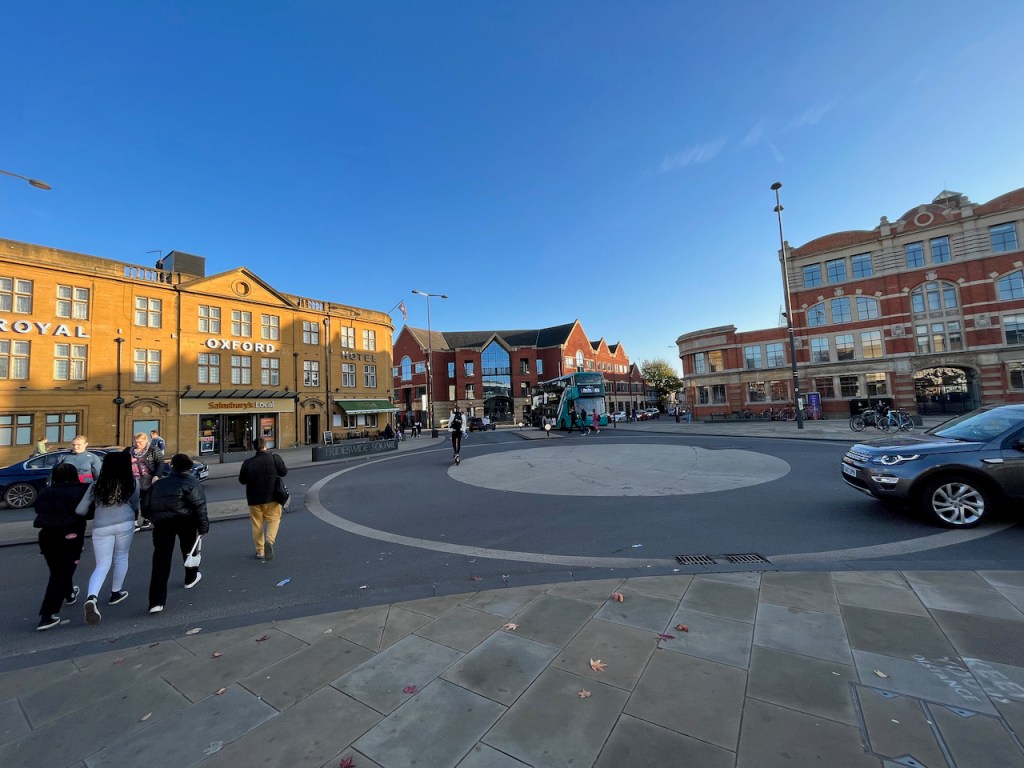 A section of Frideswide Square in Oxford. The Royal Oxford Hotel overlooks a roundabout, off which there are 3 wide roads. Pedestrians are crossing these roads, which don't have any traffic lights, while cars, a bus and a person on a scooter are also attempting to navigate the space from different directions.