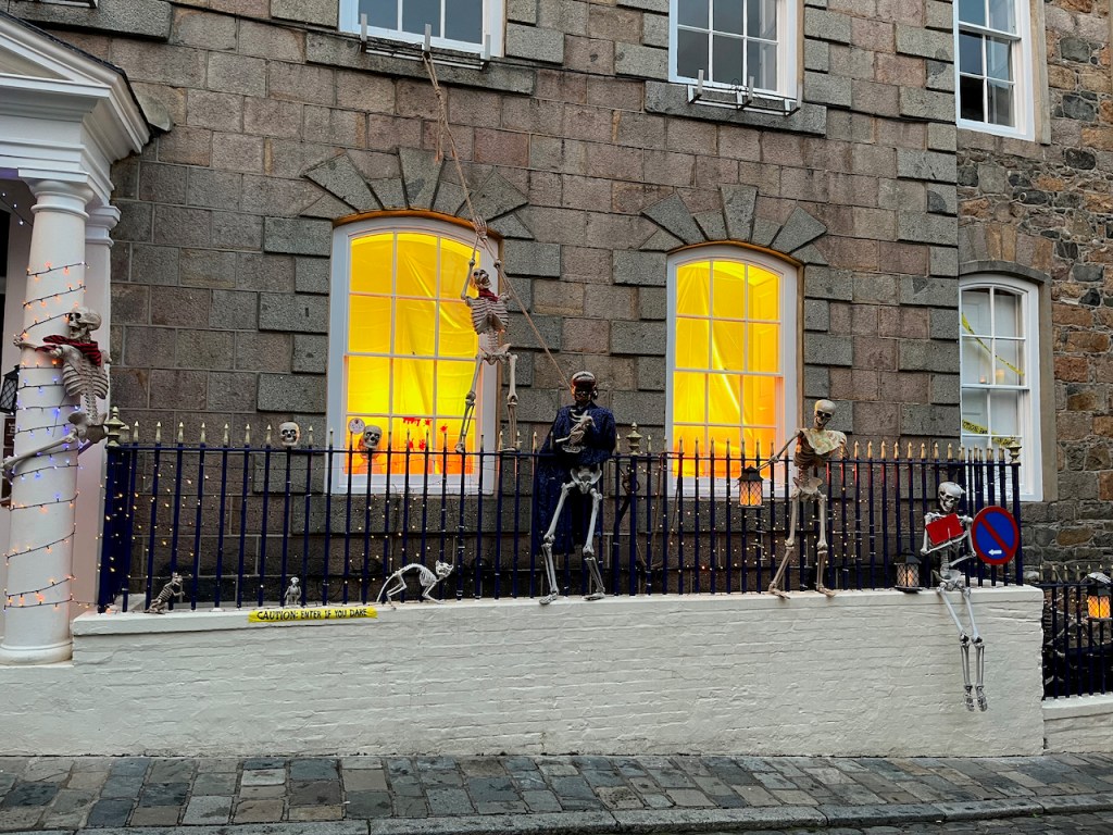 Skeletons of people and a cat in various poses attached to railings outside a large building, the windows of which are lit from the inside with colours of yellow and orange.