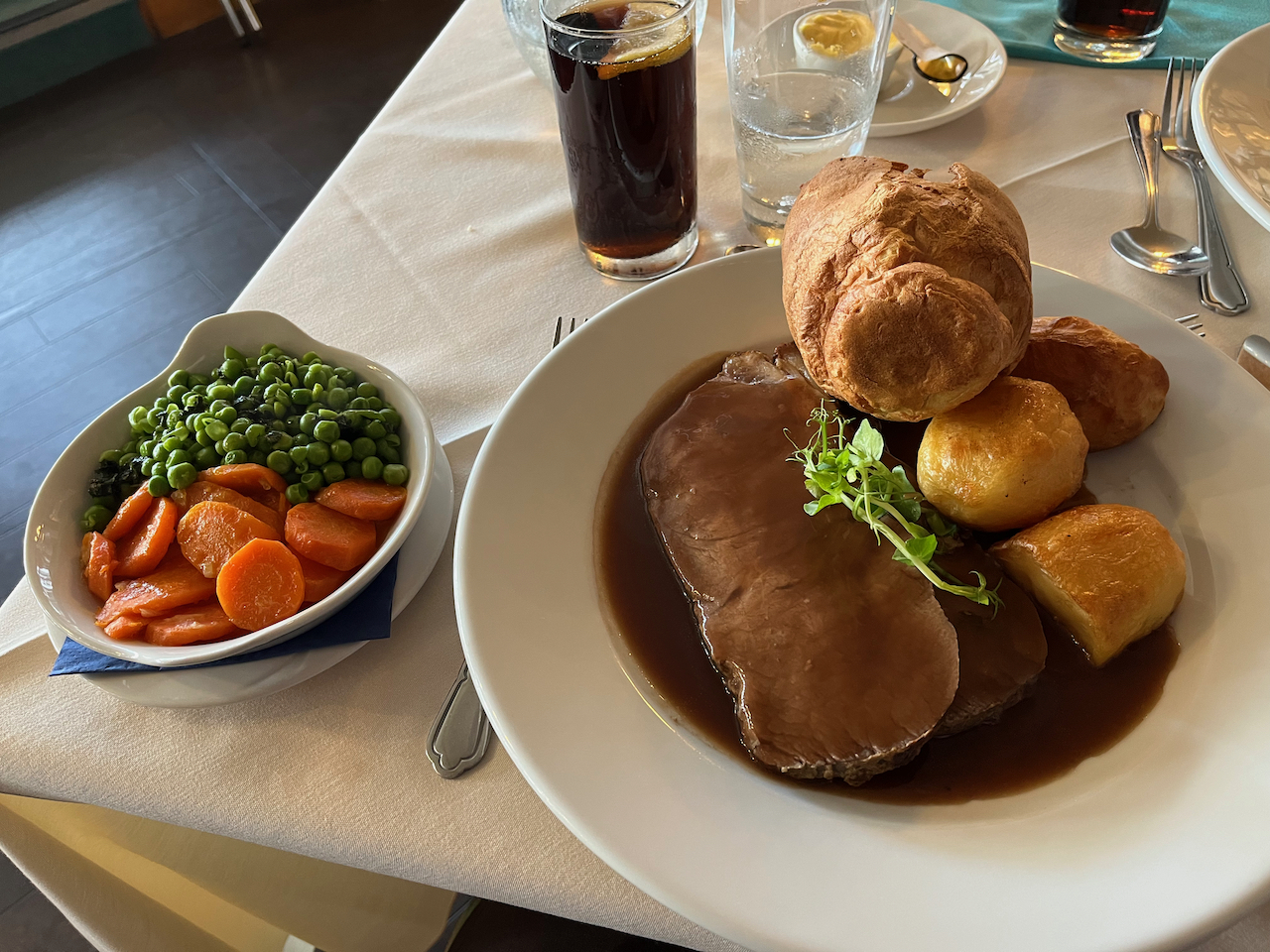 A roast dinner consisting of 2 slices of sirloin, a large Yorkshire pudding, 3 large roast potatoes and gravy, plus a separate small bowl containing carrots and peas, and drinking glasses containing Pepsi and water.