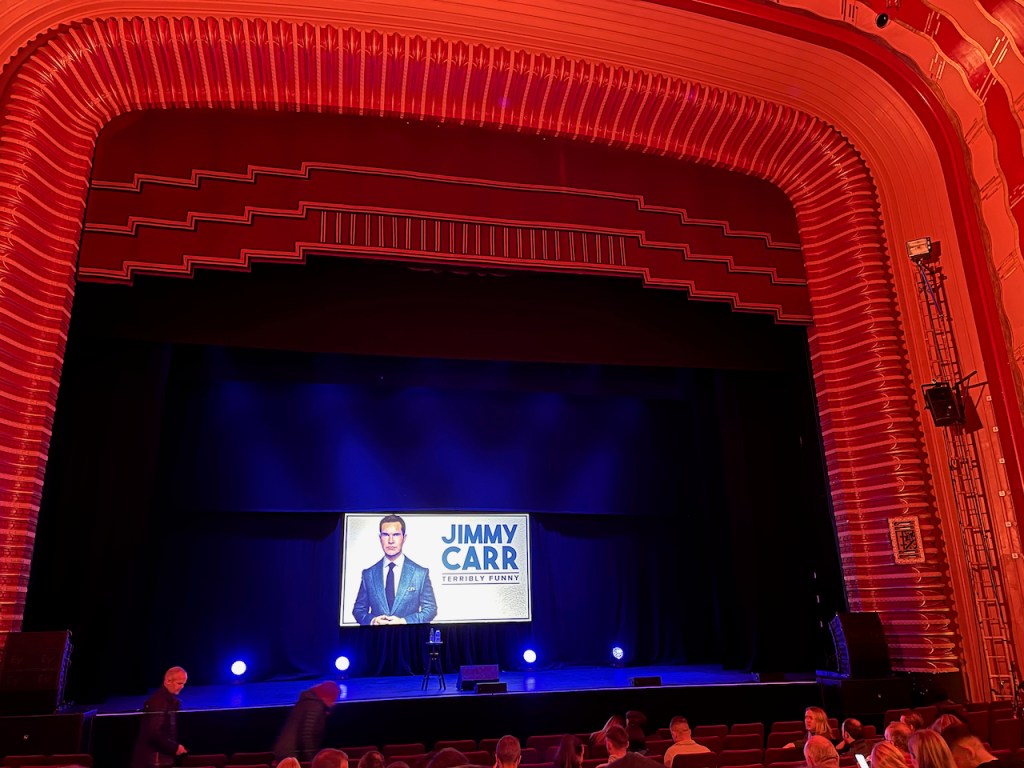 The stage for Jimmy Carr's show at the New Oxford Theatre, which has a screen at the back with a picture of Jimmy against a white background, next to his name and the title of his show, Terribly Funny.