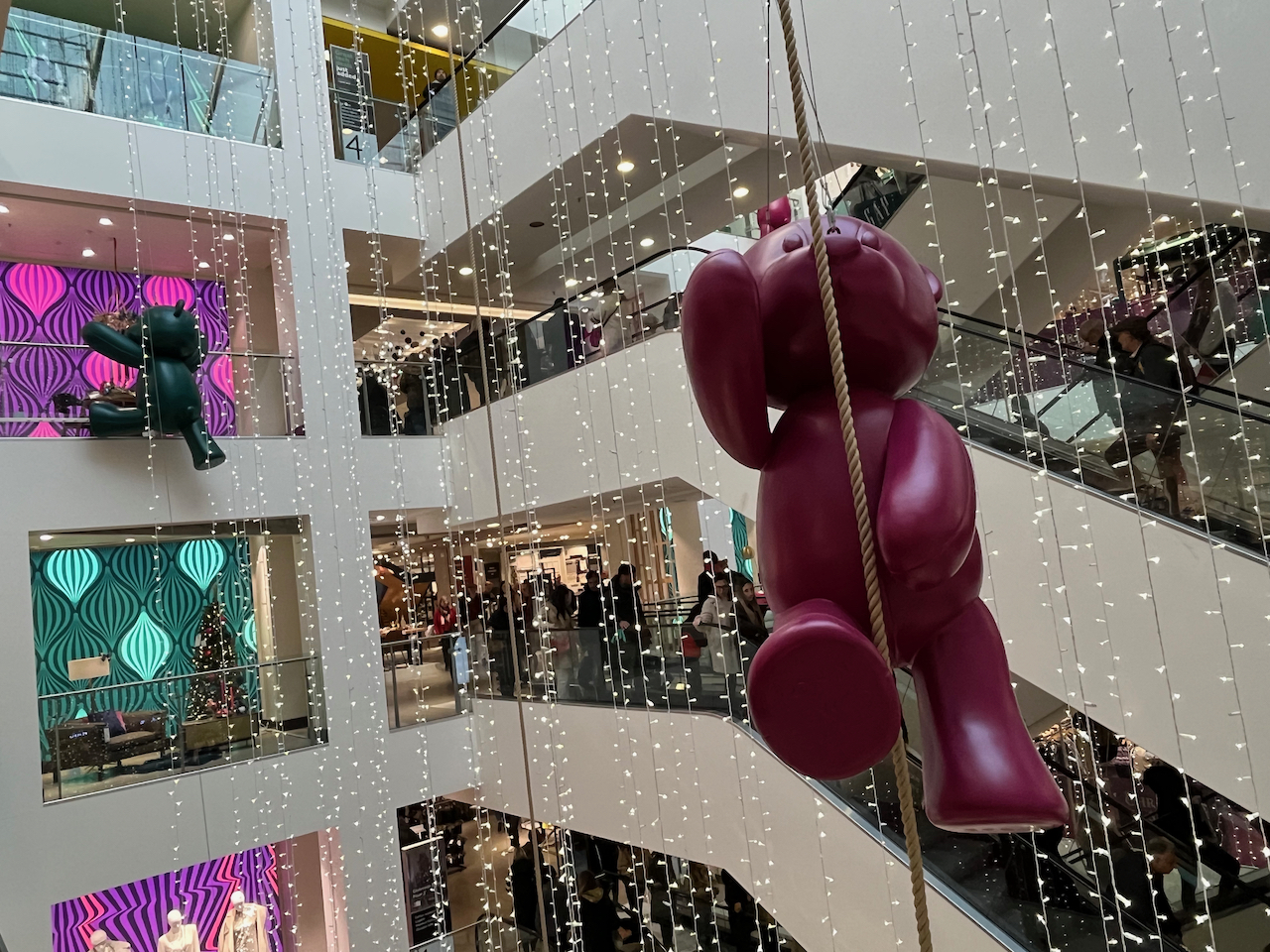 In the tall open space between the escalators and the floors of the John Lewis store in Oxford Street, a large pink model of a teddy bear appears to be climbing on a long rope suspended from the roof, while in the background a black bear is trying to climb over the railing of the nearest floor. Long strings of festive lights are also hanging down around them.