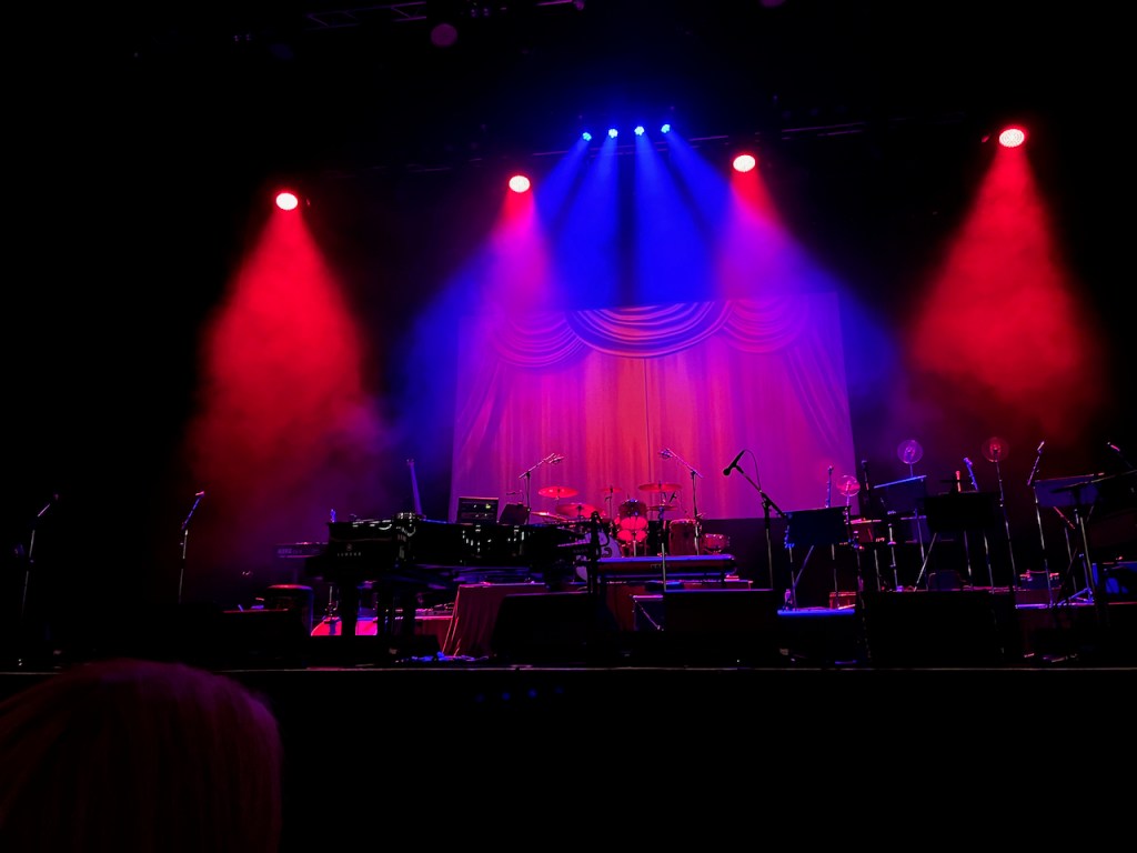 The stage before a concert by Jools Holland, featuring his piano and the many instruments played by his backing band. Blue and red lights shine down on the stage from above, and the back screen shows a closed red curtain.