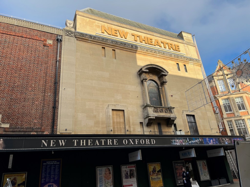 The New Theatre in Oxford, a large building with an arched window in the middle of the wall above the entrance, and the words New Theatre in big letters above the window.