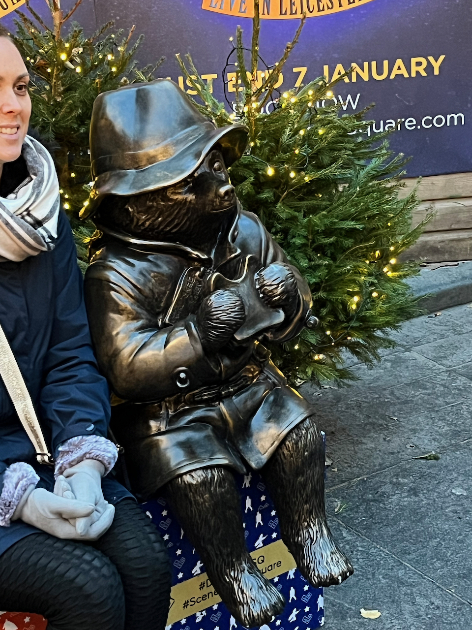 A bronze statue of Paddington Bear sitting down and eating a sandwich, in front of a green Christmas tree decorated with lights.