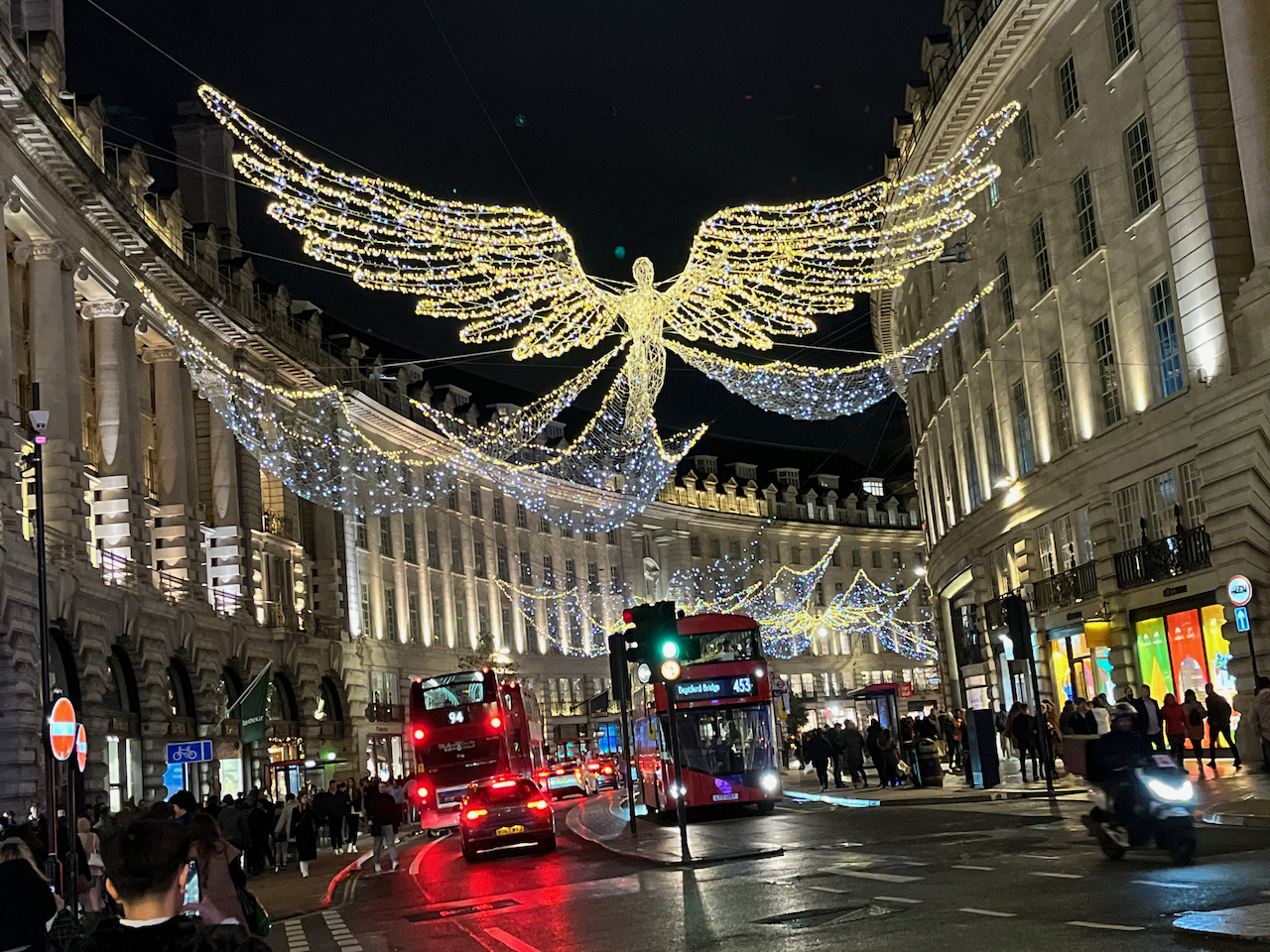 Hanging over Regent Street, and covered with glittering lights, is a figure of a person with huge wings held out at each side, and 3 large tail fins spread out behind them. Similar figures can be seen in the distance as the street curves to the right in the distance.