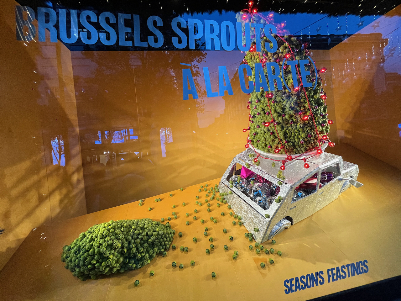 A Christmas window display at Selfridges. A silver car full of shiny Christmas decorations has a huge tower of Brussel sprouts on its roof, mixed with a few colourful baubles. There is also a big pile of sprouts behind it, and a few others scattered around, as if a lot of them have fallen off while the car's been driving along. Large text high up on the window reads Brussels Sprouts A La Carte, with brackets separating the word Car from the final two letters T and E. The words Seasons Feastings also appear at the bottom of the window.
