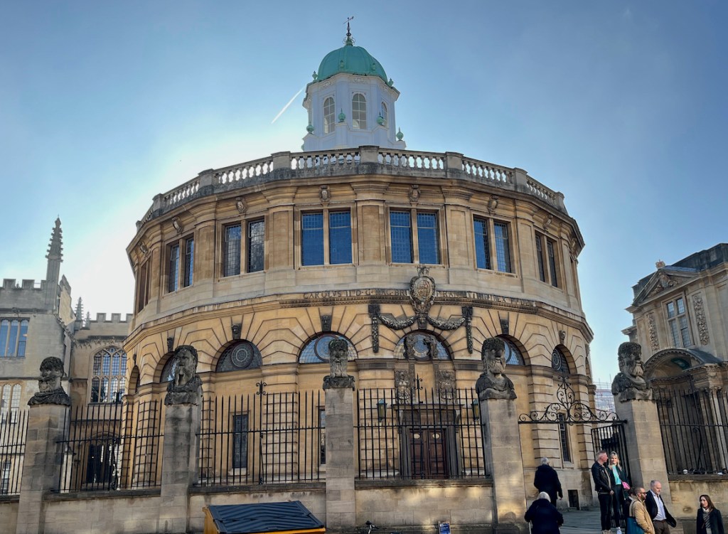 The Sheldonian Theatre, a large round 2-storey building with a small white round tower in the centre of the roof. Statues of heads stand on stone plinths in between the railings around the perimeter of the building.