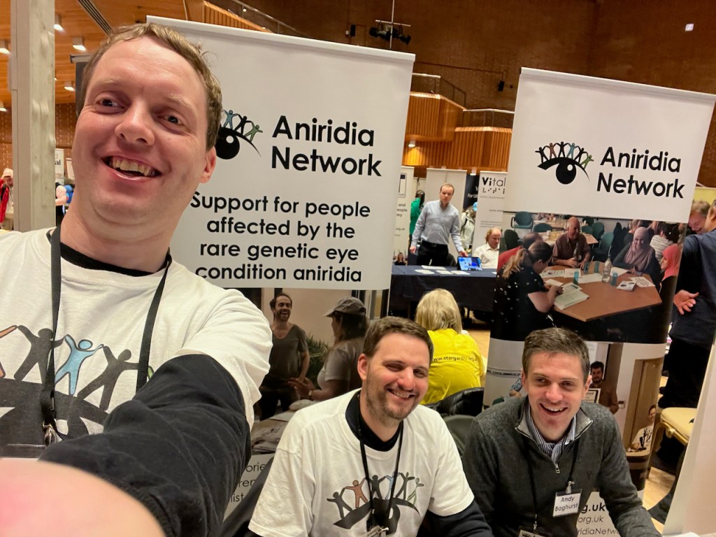 Glen, James and Andy smiling at the Aniridia Network stall at the Sight Village exhibition. Glen and James are wearing white t-shirts with the charity's logo - a large black silhouette of an eye with 5 coloured figures standing side by side on top of the eyebrow. On the tall banner behind them, below the Aniridia Network logo, text reads support for people affected by the rare genetic eye condition aniridia.