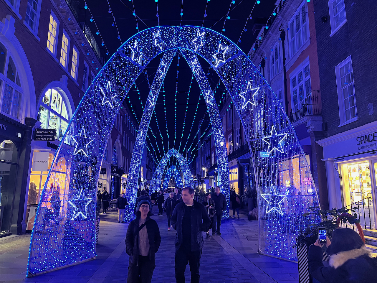 In the centre of South Molton Street, people walk through a corridor made of tall archways covered in glittery blue lights and decorated with illuminated stars, while strings of lights hang all the way along the length of the street above.