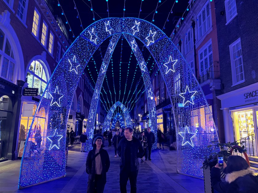 In the centre of South Molton Street, people walk through a corridor made of tall archways covered in glittery blue lights and decorated with illuminated stars, while strings of lights hang all the way along the length of the street above.