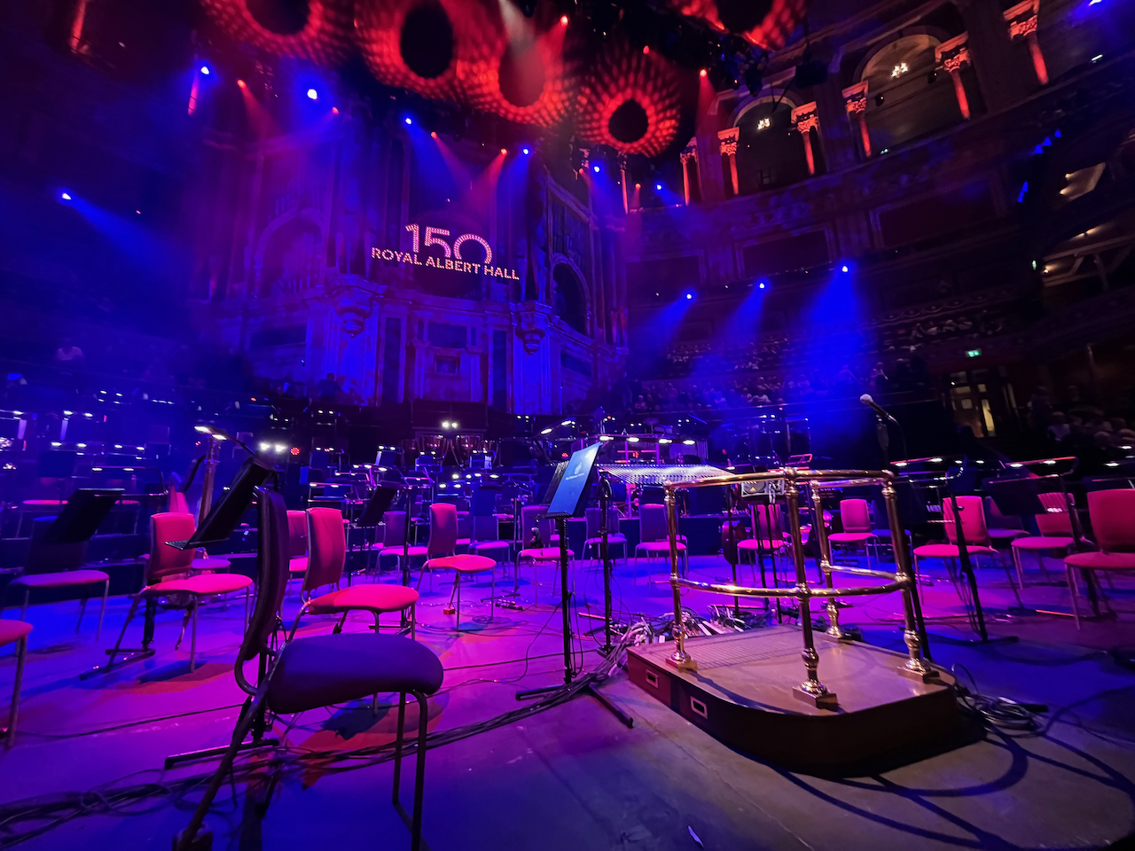 The central section of the stage at the Royal Albert Hall for the Symphonic Queen concert, lit up in blue, with chairs set up for the orchestra and a podium surrounded by metal rails for the conductor. High on the back wall, lights spell out the number 150 above the words Royal Albert Hall, marking the venue's big anniversary.