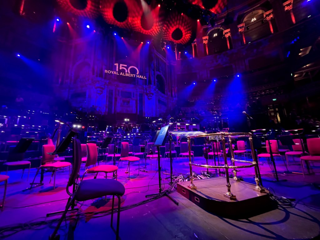 The central section of the stage at the Royal Albert Hall for the Symphonic Queen concert, lit up in blue, with chairs set up for the orchestra and a podium surrounded by metal rails for the conductor. High on the back wall, lights spell out the number 150 above the words Royal Albert Hall, marking the venue's big anniversary.