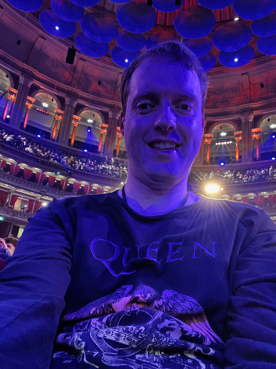 A selfie of me smiling in the Royal Albert Hall auditorium. I'm wearing a black jumper that has the word Queen above a colourful version of the band's coat of arms design.